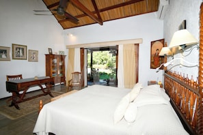 A bedroom with a large bed, wooden headboard, a desk, and a chair. There is a window with curtains, a door leading outside, and a wall-mounted lamp.