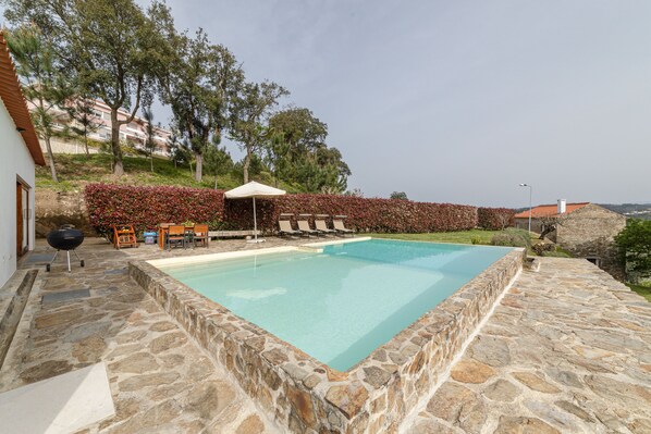Ein Pool mit Steinboden, umgeben von einer Hecke, und eine Grillgelegenheit.