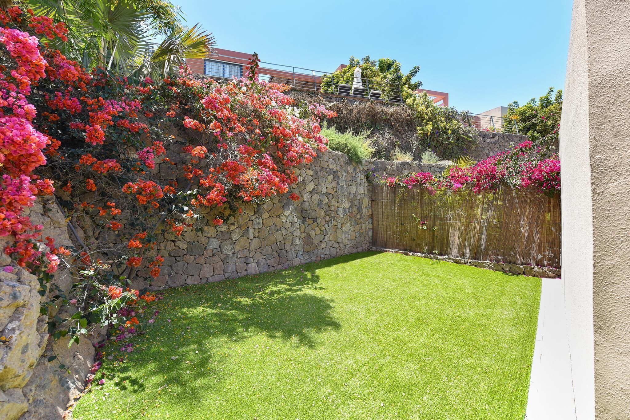 A well-maintained garden with a stone wall, lush green lawn, and vibrant flowering plants.