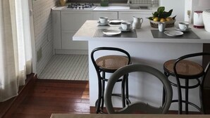 Una pequeña cocina con armarios blancos, un refrigerador blanco, una ventana con cortinas y un comedor con dos sillas.