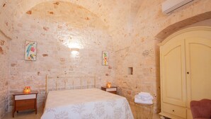 A bedroom with a stone wall, a bed with a floral bedspread, a wooden nightstand, a small table with a lamp, and a wardrobe.