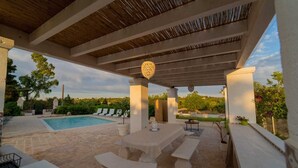An outdoor pool area with a covered patio, stone flooring, and a view of the surrounding landscape.