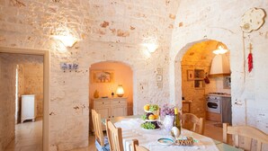 A rustic dining area with a stone wall, wooden furniture, and a kitchen visible through an archway.