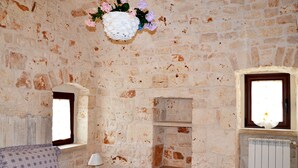 A rustic stone-walled room with a sofa, a small table with a lamp, a flower vase, and a window with a view.