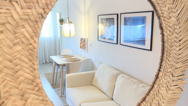 A circular mirror with a woven frame reflecting a room with a white sofa, a small table, and two framed pictures on the wall. - Santa Cruz Ocean Apartment - II (Santa Cruz)