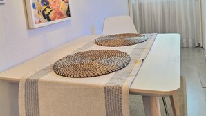 A room with a long white table, two woven placemats, a hanging lamp, a framed artwork, and a window with sheer curtains. - Santa Cruz Ocean Apartment - II (Santa Cruz)