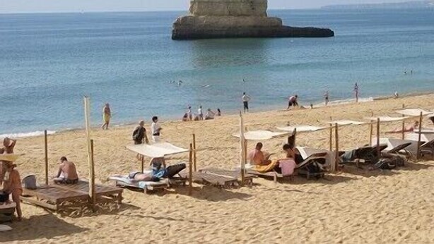Una platja amb gandules i ombrel·les, una gran formació rocosa a l'aigua, i gent nedant i relaxant-se.