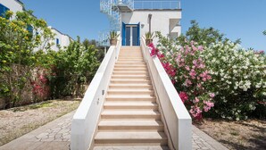 Eine Treppe führt zu einem Gebäude mit einer blauen Tür, umgeben von blühenden Pflanzen und einem Kiesweg.