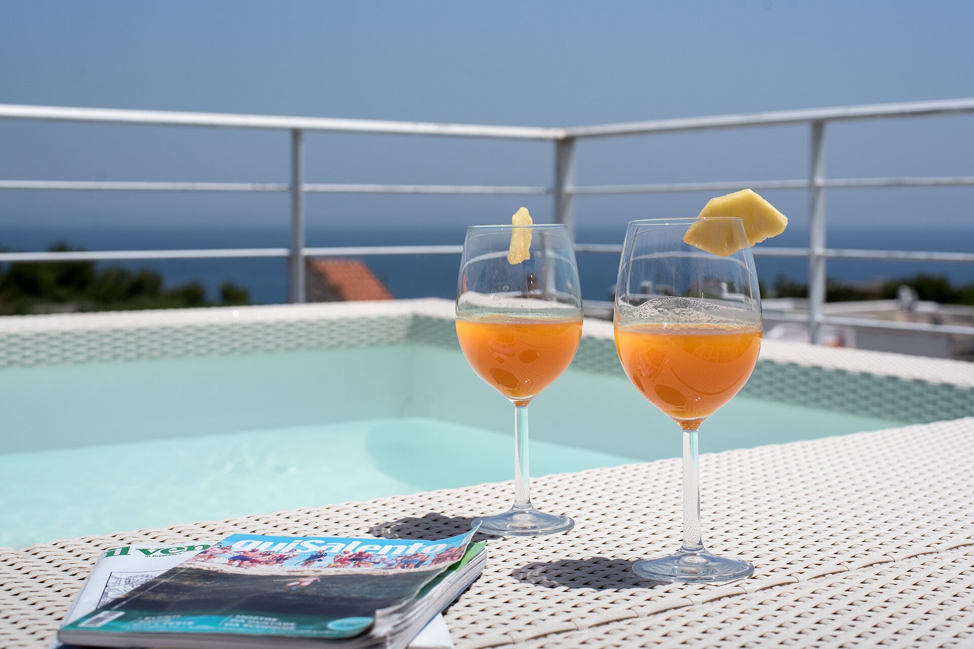 Dos vasos de jugo de naranja sobre una mesa de mimbre blanco, con una revista, con vista a una piscina y al mar.