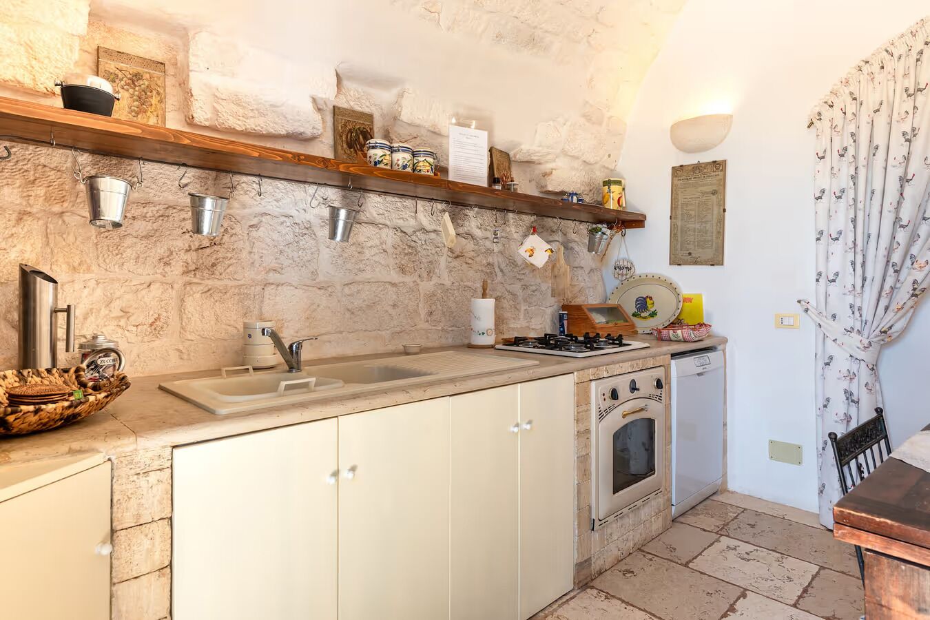 A compact kitchen with a marble countertop, a built-in oven, and a sink. There are hanging pots and pans, and a wooden shelf above the sink.