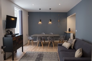 A modern living room with a dark blue wall, a wooden dining table with clear chairs, a dark sofa with cushions, and a mounted TV.