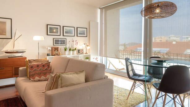 En moderne stue med sofa, sofabord i glas og stole. Væggene er dekoreret med billeder, og et stort vindue giver udsigt til bygninger.