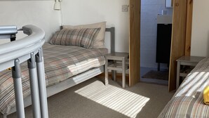 A small, compact room with two beds, a desk, and a chair. There is a door leading to a bathroom with a shower and a toilet.