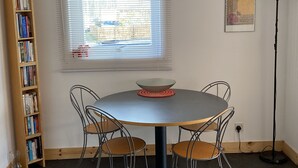 A room with a round table, four chairs, a bookshelf, a window with blinds, a floor lamp, and a wall-mounted picture.