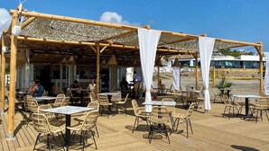 An outdoor restaurant with wooden tables and wicker chairs, a covered seating area, and a view of the beach. - A modern penthouse (110 m2) with sea and pool view, 3 bedrooms and 2 baths (Alcaidesa, San Roque, Sotogrande)