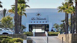 The entrance to La Hacienda with a modern design, featuring a large sign, palm trees, and a stone pathway leading to the entrance. - A modern penthouse (110 m2) with sea and pool view, 3 bedrooms and 2 baths (Alcaidesa, San Roque, Sotogrande)