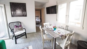 Zona pranzo con un tavolo apparecchiato per due, una sedia nera, una sedia bianca e una sedia in vimini nero. Si nota un quadro appeso alla parete, un televisore montato sopra il tavolo e una finestra con infissi bianchi.