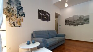 Un salon avec un canapé bleu, un pouf blanc, une petite table ronde sur laquelle se trouve une tasse bleue, et un mur orné d’œuvres d’art abstraites et représentant des paysages urbains.