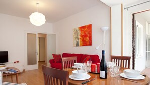 Een moderne woonkamer met een rode bank, een eettafel gedekt met borden, glazen en een fles wijn. Er is een televisie, een boekenkast en een schilderij aan de muur.