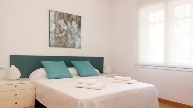 Een netjes opgemaakt slaapkamer met een bed, kussens, een nachtkastje, een lamp en een schilderij aan de muur.