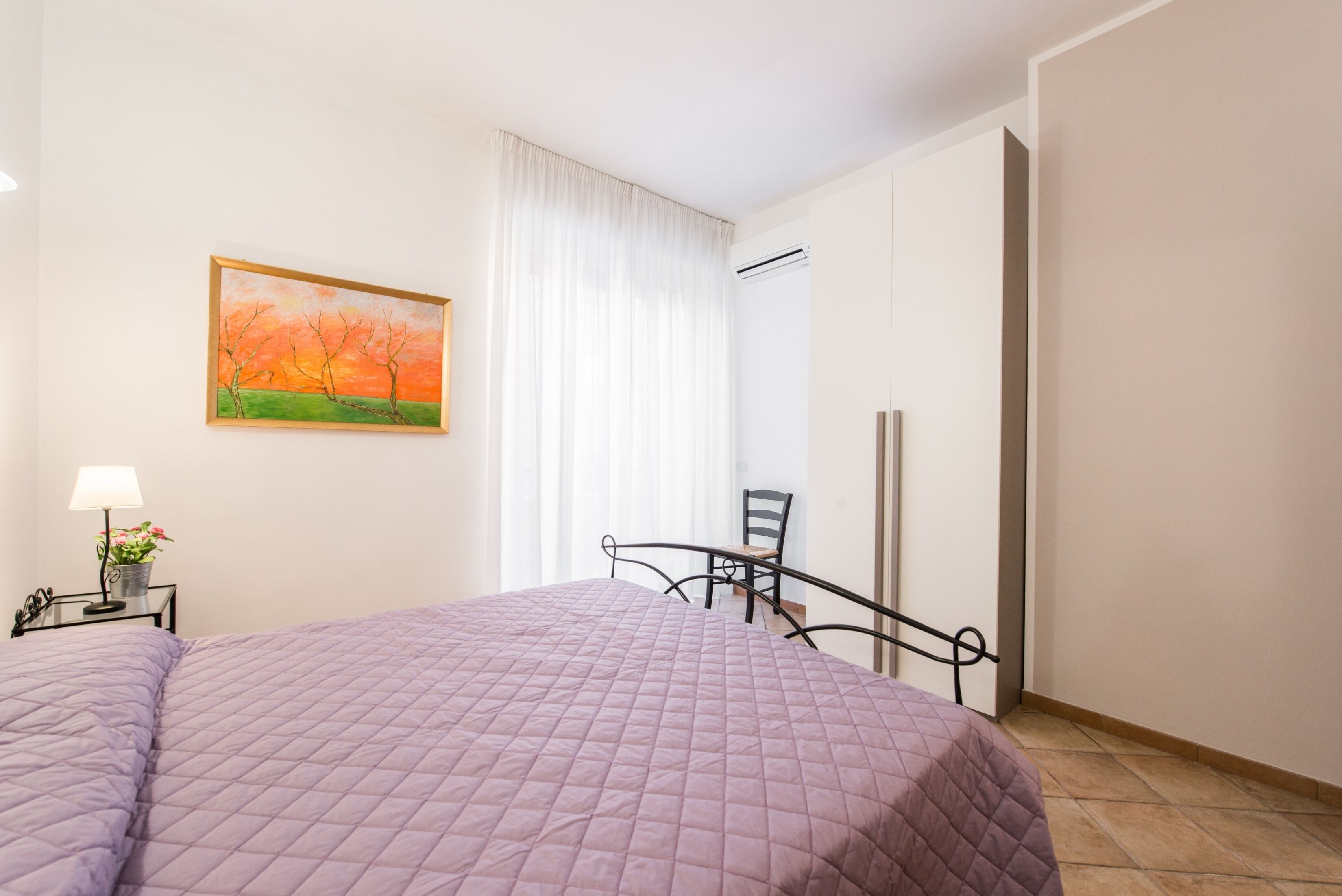A bedroom with a bed, a chair, a nightstand, a lamp, and a painting on the wall.