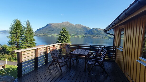 Une terrasse en bois avec une table et des chaises, d’où l’on a vue sur un lac et des montagnes.