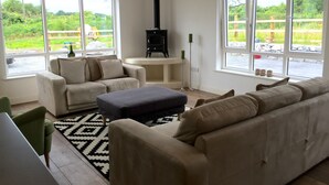 Un salon avec un canapé beige, un pouf bleu et un fauteuil blanc. On y trouve une cheminée noire, une table basse blanche et un lampadaire noir. De grandes fenêtres offrent une vue sur un jardin et un mur en pierre.