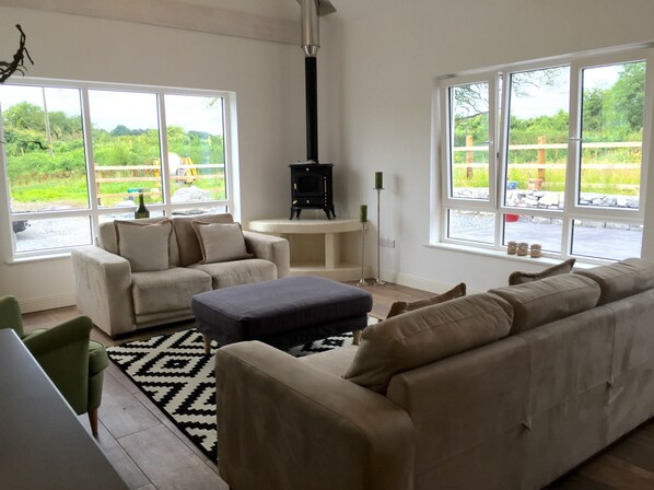 Una sala de estar con un sofá color beige, un reposapiés azul y un sillón blanco. Hay una chimenea negra, una mesa de centro blanca y una lámpara de pie negra. Amplias ventanas ofrecen vistas a un jardín y un muro de piedra.