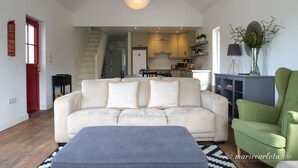 Un vaste salon avec un canapé blanc, un fauteuil vert et un pouf gris. Un escalier mène à l’étage supérieur, et on aperçoit une cuisine au fond.