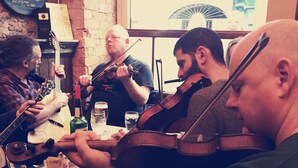 Un groupe de musiciens jouant d’instruments à cordes dans un cadre rustique, avec des murs en briques et une fenêtre en arrière-plan.