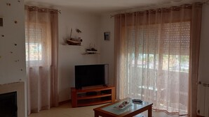 Un salon avec une cheminée, un téléviseur posé sur un meuble en bois, une table avec une télécommande et une fenêtre ornée de rideaux légers.