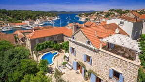 A stone building with a swimming pool, red-tiled roofs, and a marina in the background.