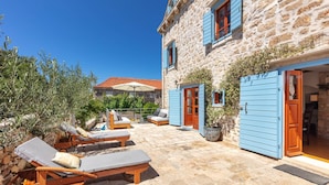 A stone building with blue shutters, a patio with lounge chairs, and a red-tiled roof in the background.