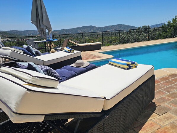 Un área de descanso junto a la piscina con sillones reclinables, una mesa con una botella y vistas a las montañas.