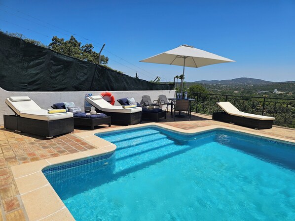 Una piscina con sillas de descanso y una sombrilla, rodeada por un muro de piedra y vistas al campo.
