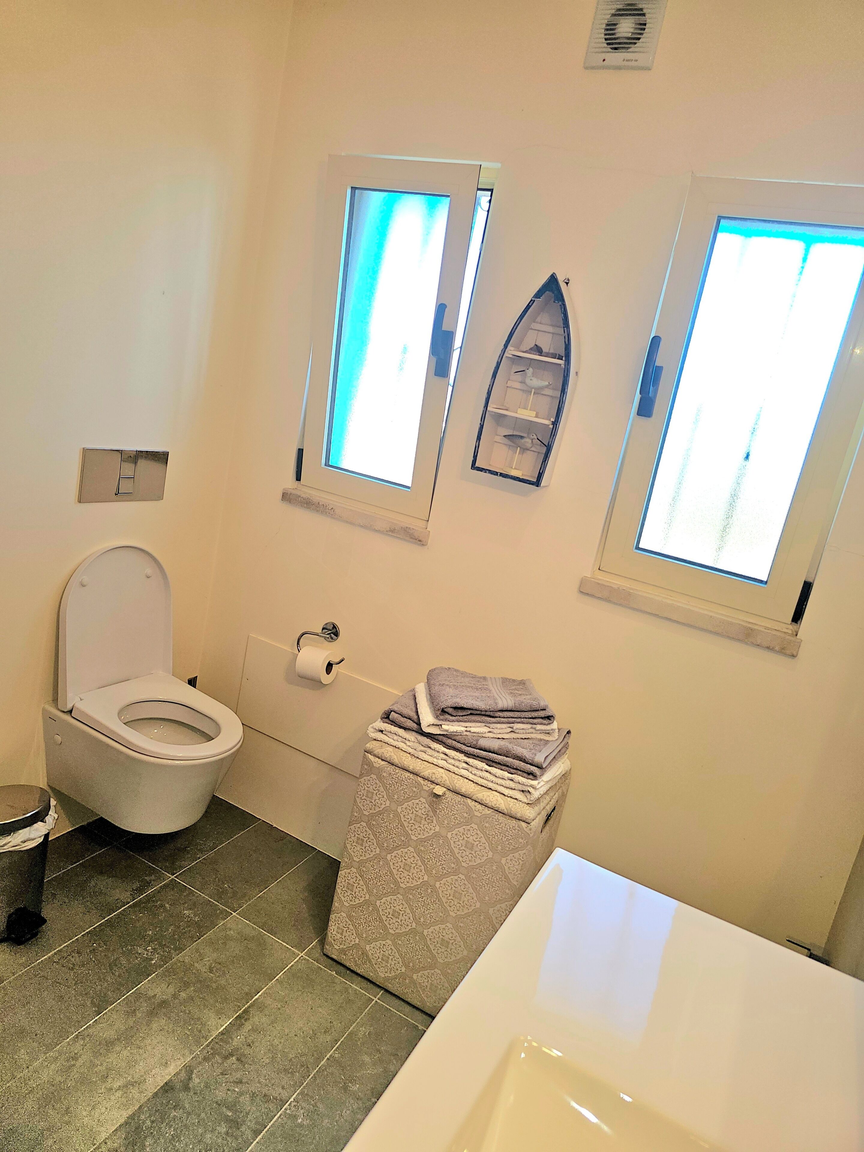 Une salle de bain comprenant des toilettes, un lavabo, une pile de serviettes et deux fenêtres.