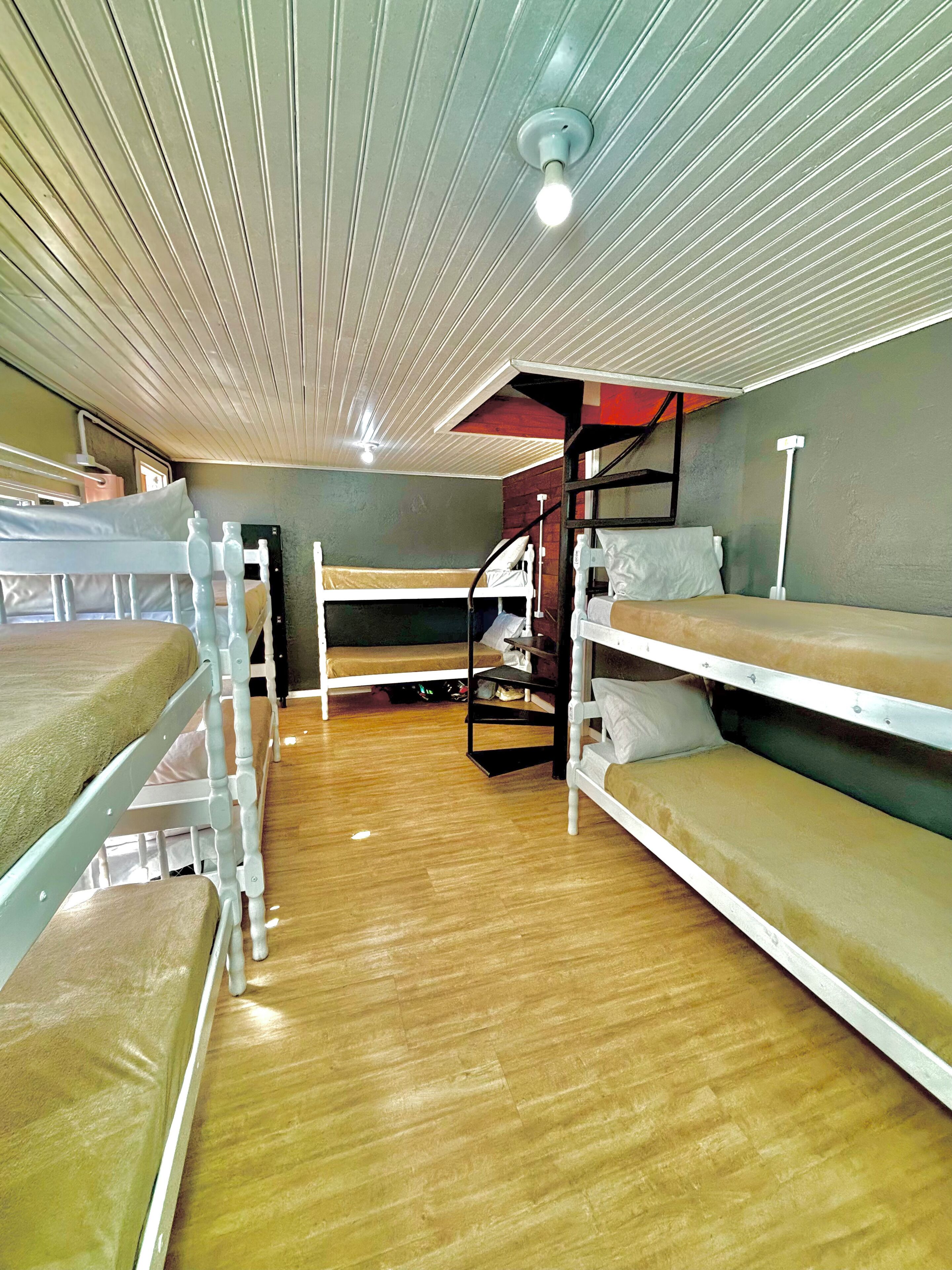 A dormitory room with bunk beds, wooden flooring, and a ceiling with linear lighting fixtures.