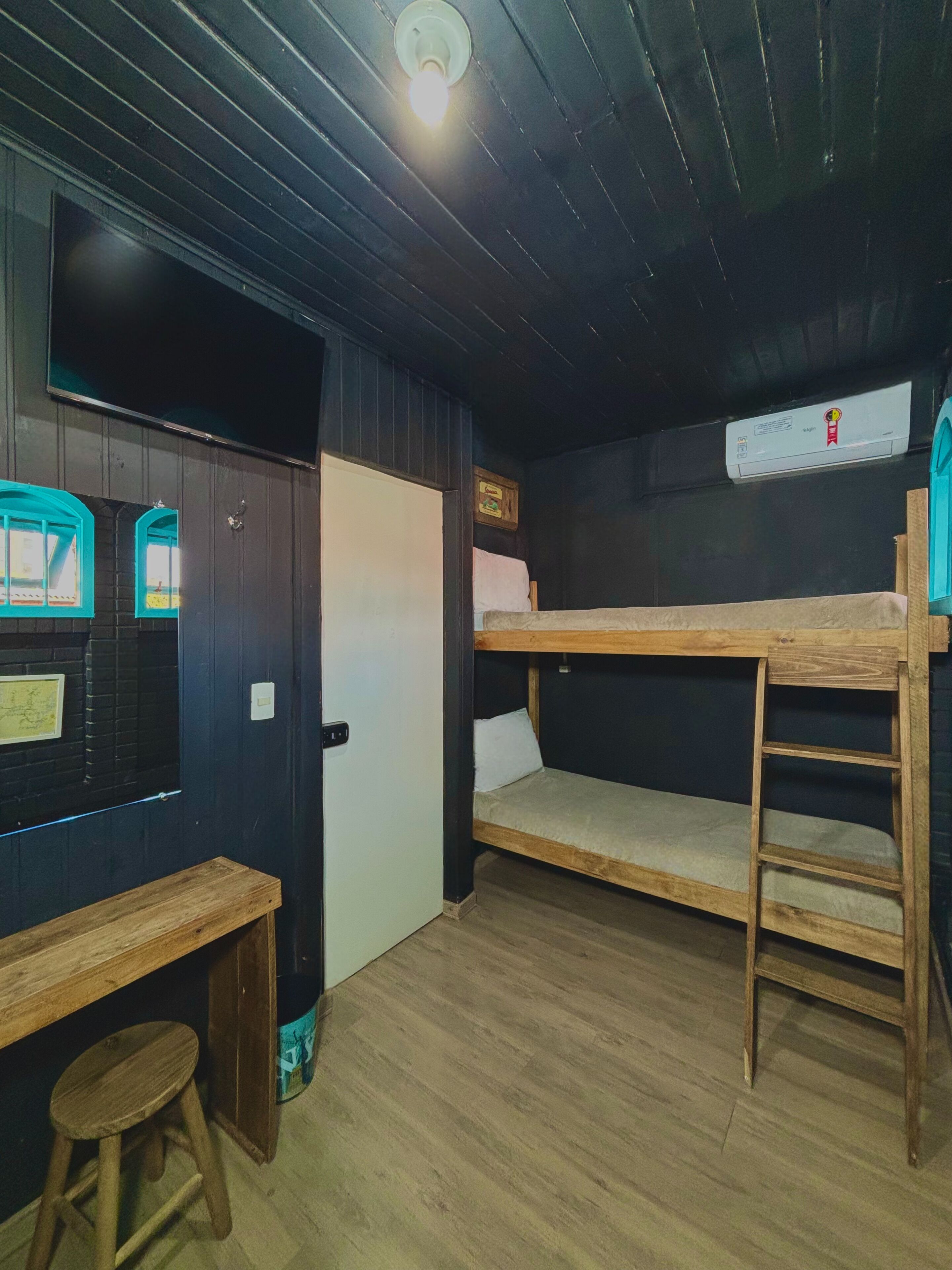 A bunk bed room with a wooden bench and a stool, a wall-mounted air conditioner, and a door with a light switch.