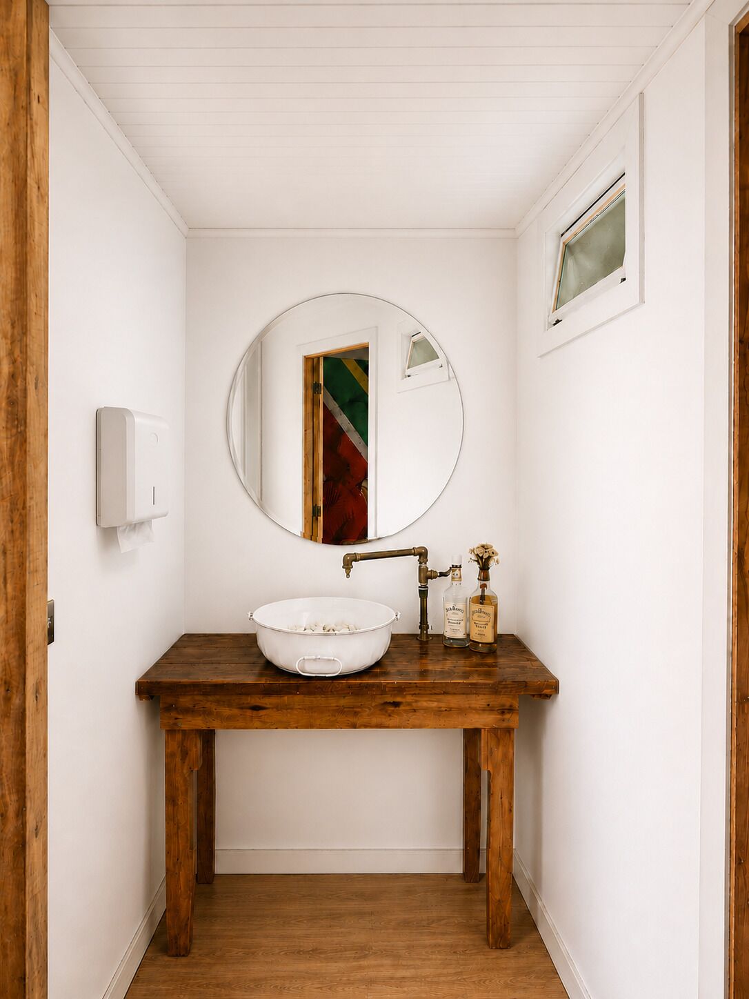 Un baño con tocador de madera, espejo redondo, lavabo blanco y una ventana pequeña.