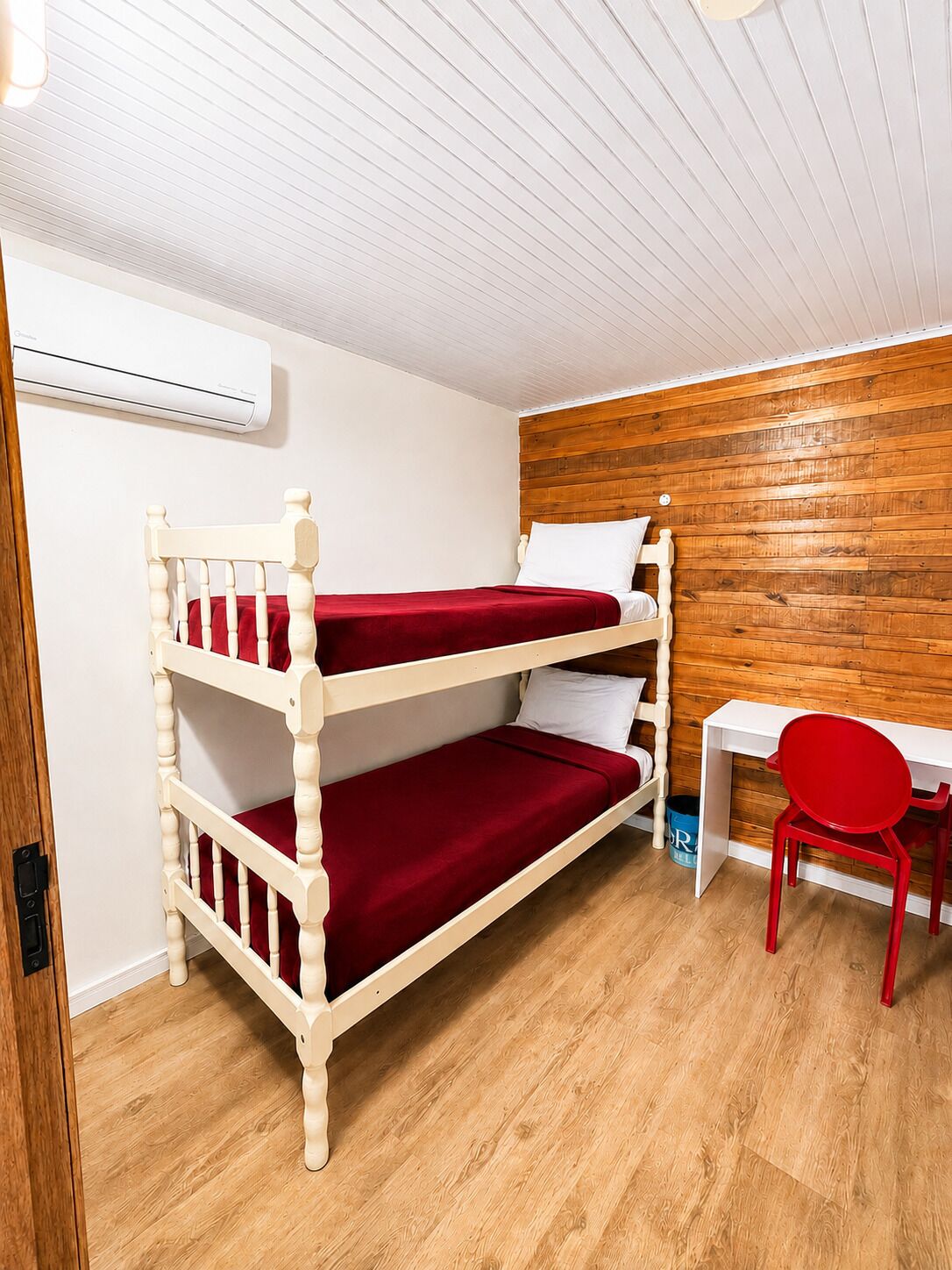 A bunk bed with red bedding, a white desk, a red chair, and a wooden wall.
