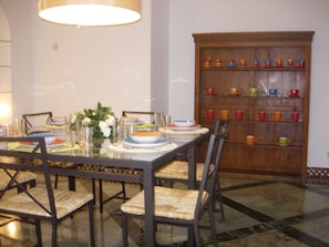 A dining area with a long table set for four, surrounded by chairs with woven seats. A wooden cabinet with colorful cups and saucers is visible in the background.