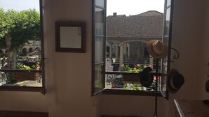 Une pièce avec un plancher en bois, un lit, une fenêtre donnant sur un immeuble et des arbres, et un chapeau accroché au mur.
