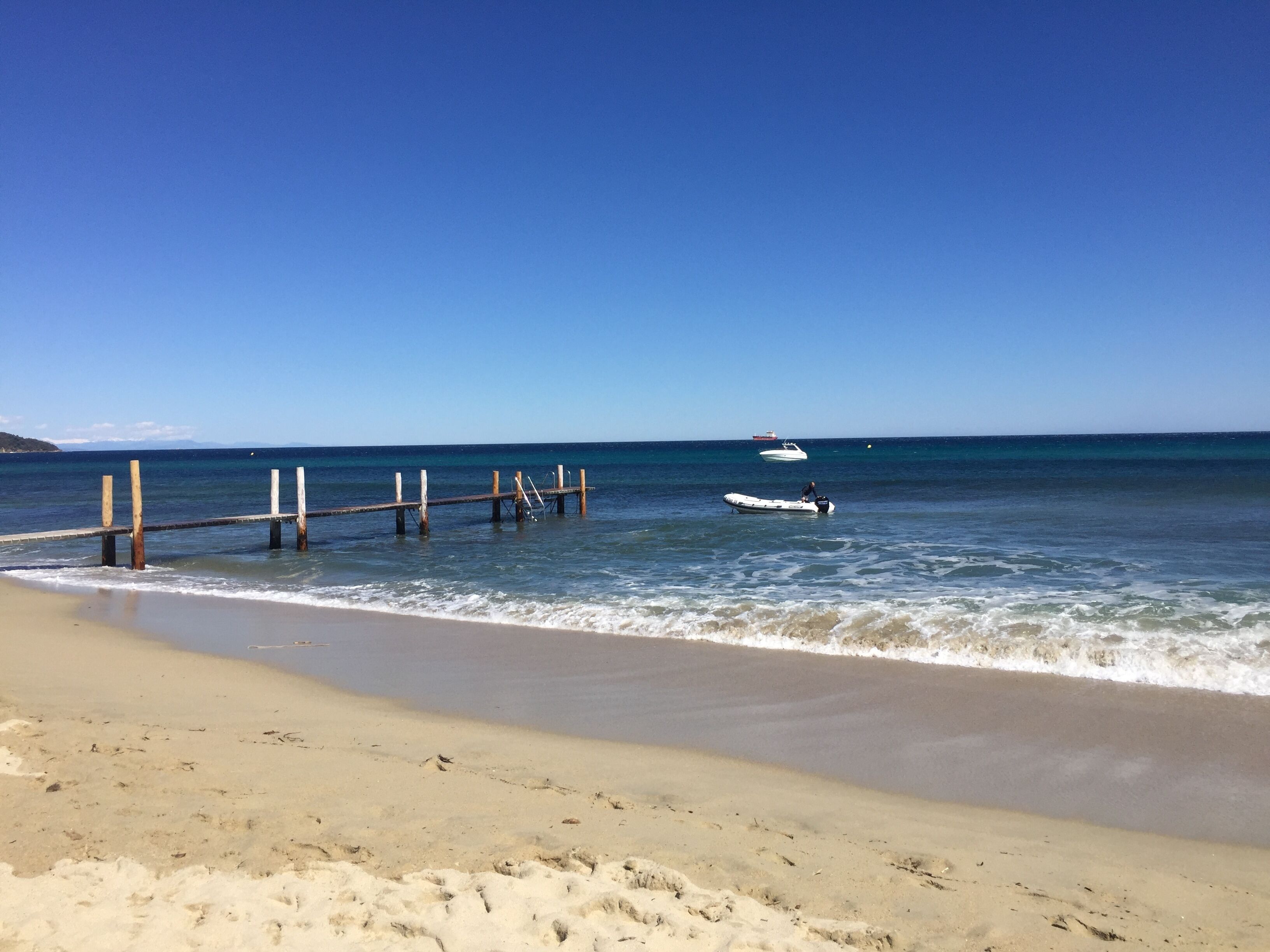 A beach with a wooden pier extending into the water, a boat anchored near the shore, and a distant ship on the horizon.