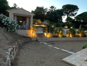 Área externa bem iluminada, com muro de pedra, degraus que levam a um edifício e diversos tipos de plantas.
