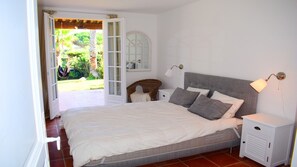 Une chambre à coucher avec un lit, des tables de chevet, une lampe et une vue sur un jardin, visible à travers une porte ouverte.