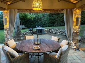 Une terrasse extérieure avec une table ronde en bois entourée de chaises en osier, un barbecue et un mur en pierre en arrière-plan.