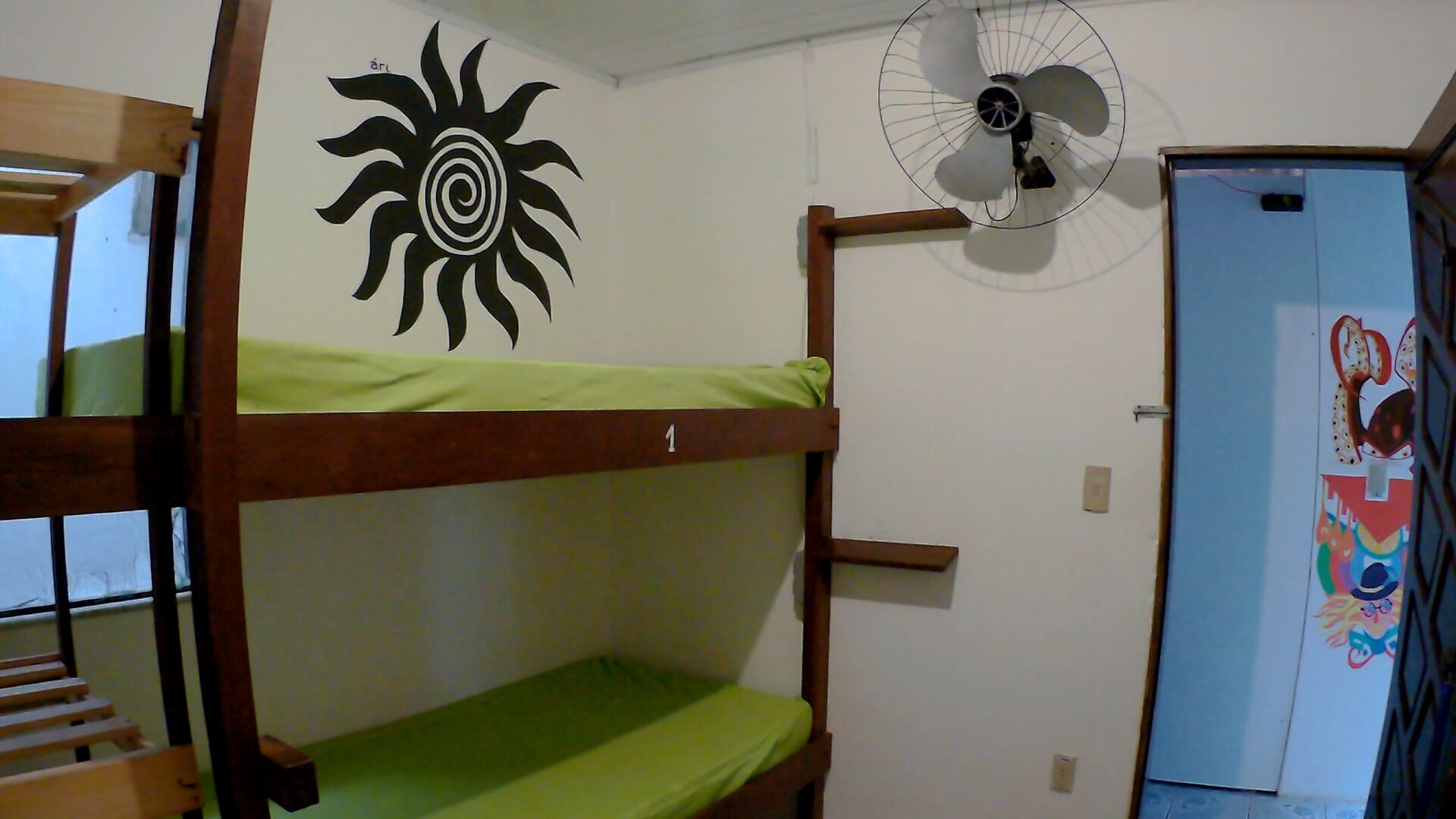 Ein Stockbett mit einer grünen Matratze, ein Wandventilator und eine Tür mit farbenfroher Gestaltung.