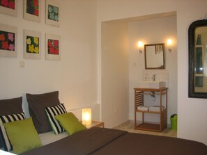 A bedroom with a bed, bedside table with a lamp, a wooden shelf with towels, a mirror, and a wall with framed floral artwork.