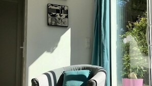 A modern living room with a grey sofa, teal cushions, and a large window with a view of greenery. - Modern studio, air conditioning, parking, wifi, terrace, 2 steps from the beach (La Ciotat)