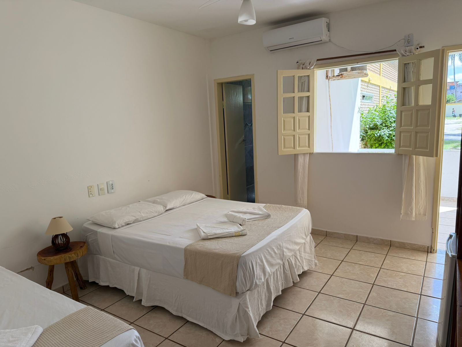 A bedroom with a bed, a bedside table with a lamp, an air conditioning unit, and a window with open shutters.
